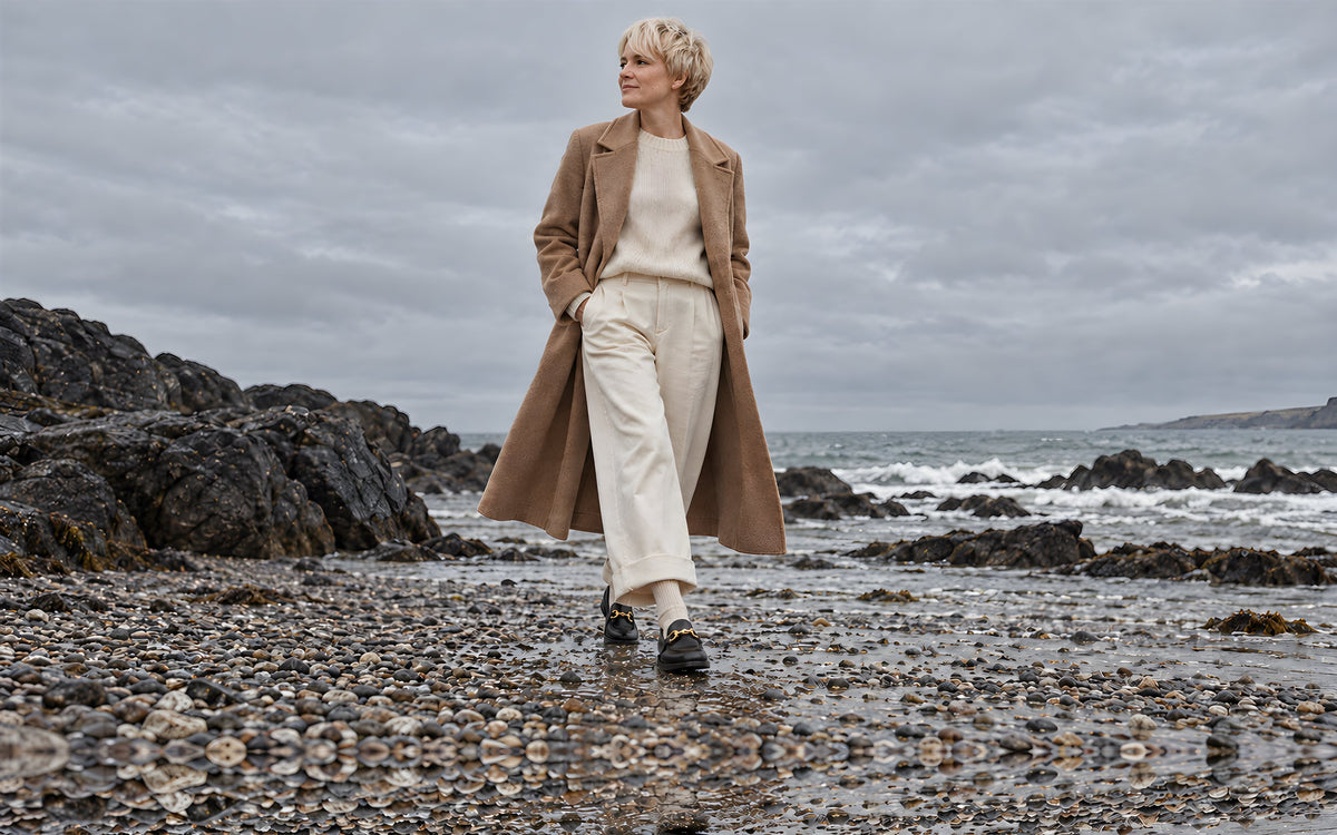 Woman in a camel coat walking on a pebble beach in black horsebit loafers